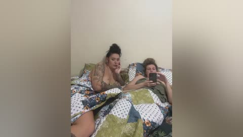 Media: Video of two people in a dimly lit room: a tattooed woman in a floral bra, and a man in a green shirt, both under patchwork quilts.