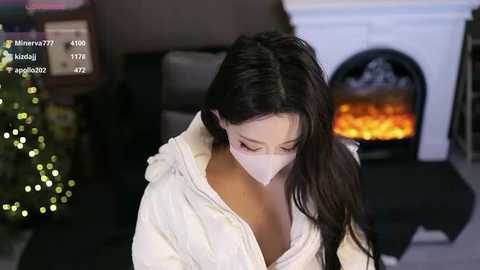 Video of a young Asian woman with long black hair, wearing a white mask and bathrobe, kneeling in a cozy living room with a lit fireplace and Christmas tree.