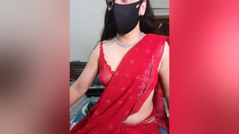Video of a woman wearing a red saree with a black mask, silver necklace, and a black face mask, seated on a bed with a blue and white blanket.