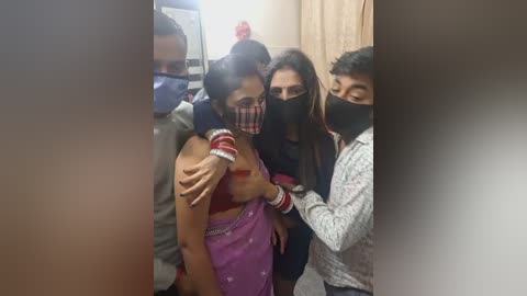 Video of a woman with a pink sari, wearing a face mask, being hugged by a man in a grey shirt and another man in a black mask, in a room with beige walls.