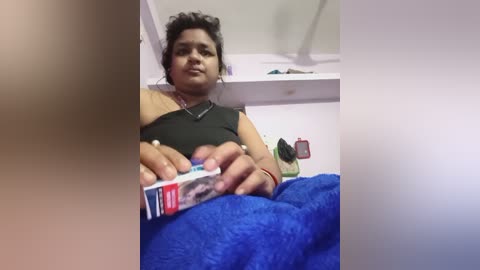 A video of a woman with dark skin, curly hair, wearing a black top and blue blanket, sitting on a bed, opening a medicine box.