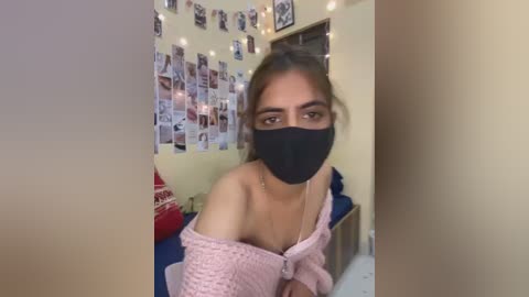 Video of a young woman with light brown skin, brown hair, wearing a black mask, off-shoulder pink knitted sweater, and a string of photos on a yellow wall in a cozy room.