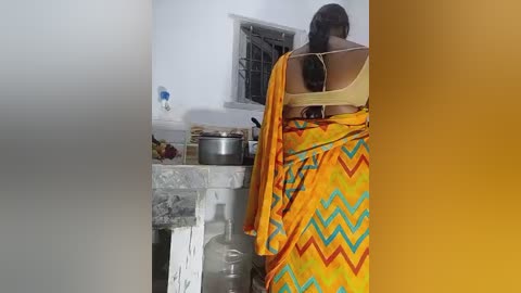 Video of a woman in a yellow sari with a zigzag pattern, standing in a rustic kitchen with a stone sink, a metal pot, and a window with bars.