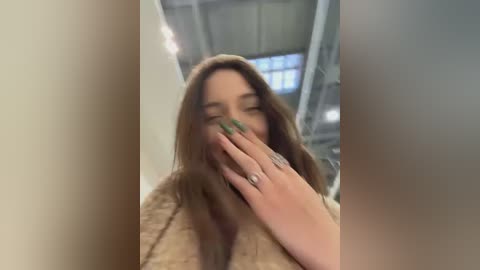 Video of a young woman with long brown hair, laughing, covering her mouth with one hand, wearing a beige sweater, in a dimly lit industrial room with high ceilings and large windows.