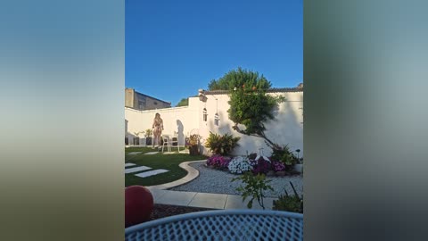 A serene video of a sunlit Mediterranean-style courtyard with a white stucco wall, lush greenery, and vibrant flower beds, framed by a glass door with a frosted edge.
