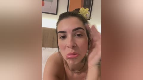 Video of a woman with light brown skin, dark hair, and a yellow flower, lying on a bed with a white sheet, looking directly at the camera with a hand on her face, partially blurred.