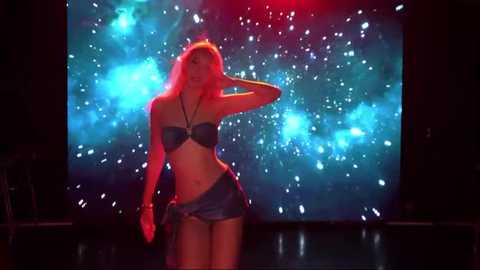 Video of a curvy, blonde, topless woman in a silver bikini, standing confidently on a stage with a vibrant, cosmic background and dramatic lighting.