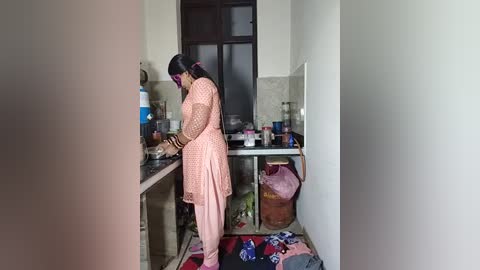Media: Video of a woman in a light pink, long-sleeved dress with a polka-dot pattern, cooking on a stove in a small, cluttered kitchen with a white countertop, tiled backsplash, and a pink bag on the floor.