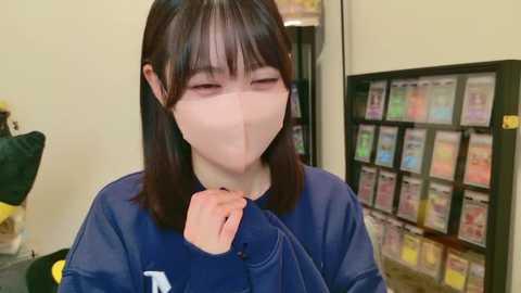 A video of a young woman with straight, shoulder-length dark hair, wearing a blue shirt and a clear face mask, sniffing her fingers. Background includes a vending machine with colorful snack options and a green object.