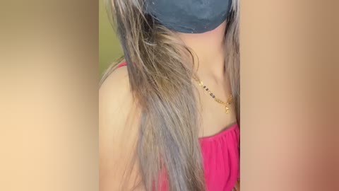 Video of a light-skinned woman with long, straight, light brown hair, wearing a black face mask, pink sleeveless top, and a gold necklace. Background is blurred, focusing on her face and neck.