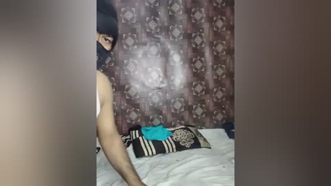 Video of a person in a black mask, wearing a white tank top, standing in a room with patterned brown wallpaper, bed with white sheets, and a blue item on the bed.