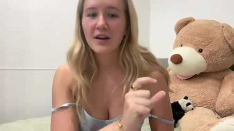 A video of a young blonde woman with fair skin, wearing a light blue bra, gesturing with her left hand, standing beside a large teddy bear in a simple, beige room.