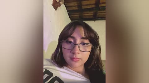 A video of a young, light-skinned woman with shoulder-length brown hair, wearing glasses and a white T-shirt with a black design. She has a neutral expression, and the background shows a wooden ceiling and white wall.