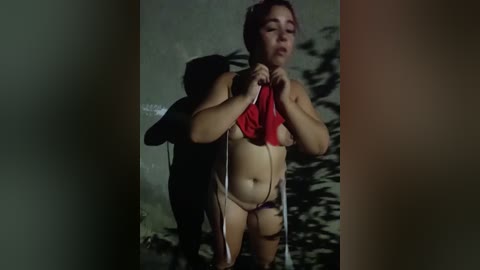 A video of a nude woman with a red shirt covering her breasts, standing in a dimly lit, shadowy room with a wall of green foliage in the background.