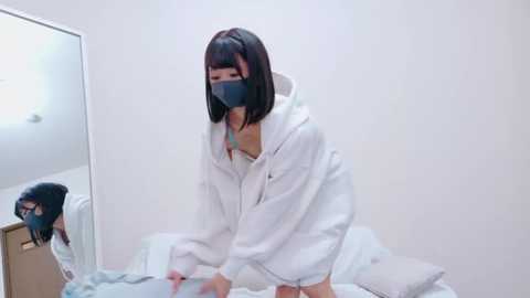 Video of an East Asian woman with a slender build, wearing a white hoodie and a black face mask, kneeling on a bed. The room is minimalistic with white walls and a mirror.