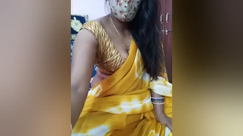 Video of a woman in a yellow sari with gold accents, wearing a floral face mask and gold jewelry, sitting in a domestic setting with a blurred background.