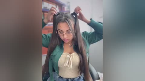 Video of a young woman with long, straight black hair, wearing a green cardigan, beige top, and glasses, adjusting her hair in a casual indoor setting with blurred shelves in the background.