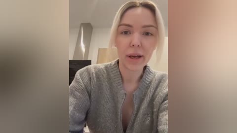 Video of a blonde woman in a gray cardigan, with a neutral expression, standing in a modern, softly lit room with beige walls and a black lamp.