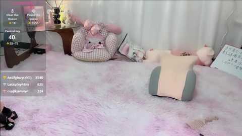 Video of a cozy, pastel-themed room with a plush, white furry rug. A gray and beige massage chair sits on the rug, surrounded by pink and white pillows and a checkered blanket.