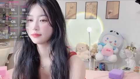 A video of a young East Asian woman with long black hair, light skin, and red lipstick, standing in a cozy room with plush toys, framed artwork, and a white lamp.