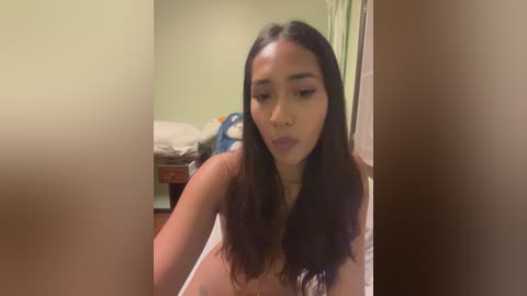 Video of a young, light-skinned woman with long, straight brown hair, wearing a white tank top, in a dimly lit bedroom with a blue bag and unmade bed in the background.