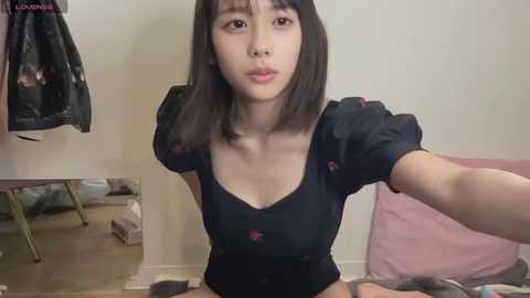 Media: Video of a young East Asian woman with straight black hair, wearing a black floral dress, sitting on a bed in a cluttered bedroom.