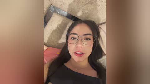 Video of a young Asian woman with long black hair, wearing black-framed glasses and a black top, leaning against a stone wall with a large, metallic object behind her.