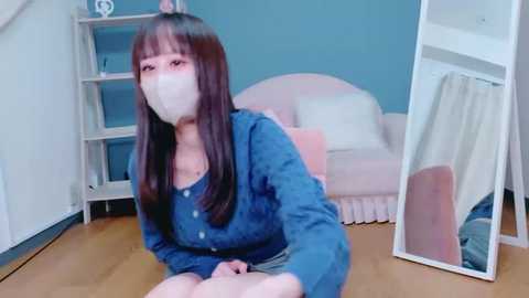 Media: Video of an Asian woman with long black hair, wearing a blue sweater, face mask, and sitting on a wooden floor in a cozy, pastel-colored room with a pink sofa, white curtains, and shelves.