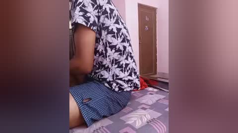 Video of a person kneeling on a patterned carpet, wearing a black floral shirt and blue checkered shorts, in a room with pink walls and a wooden door.