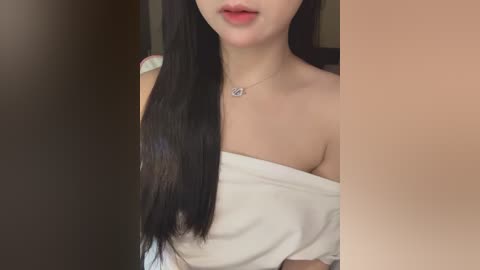 Video of a young Asian woman with long, straight black hair, fair skin, and light makeup, wearing a white off-shoulder top and a delicate necklace, in a softly lit room.