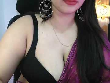 Video of a woman with fair skin, long black hair, wearing a black bra, a purple dupatta, and large silver hoop earrings. Background features a warm, softly lit room with a wooden shelf.