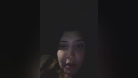 A dimly lit video of a woman with fair skin, dark hair, and a pensive expression, partially obscured by shadows. The background is dark and indistinct.