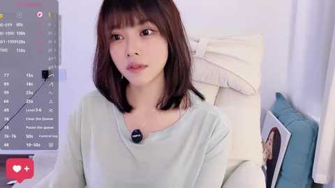 Video of an Asian woman with straight, shoulder-length brown hair, wearing a light blue long-sleeve top, sitting on a bed with white pillows and a blue cushion, against a white wall, with a TV displaying a calendar in the background.