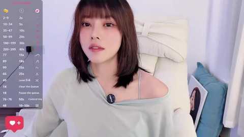 Video of an Asian woman with straight, shoulder-length brown hair, wearing a light gray off-shoulder sweater, sitting on a white leather chair in a minimalist room with a white wall and blue pillow.