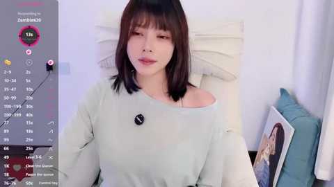 Video of an East Asian woman with shoulder-length dark hair, fair skin, and a light green off-shoulder sweater. She is sitting on a bed with white pillows and a blue cushion. The image is overlaid with a social media app interface.