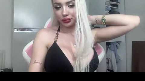 Video of a fair-skinned, platinum blonde woman with long hair, wearing a black halter top, posing in a minimalist room with a white chair and open closet.