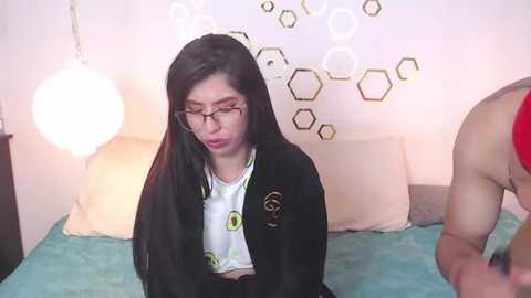 Video of a young Asian woman with long black hair, wearing glasses and a black jacket over a white T-shirt with green doodles, sitting on a bed with a teal blanket.