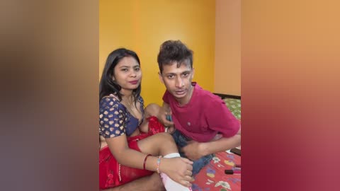 Video of a South Asian couple in a vibrant room with a baby. The woman, with long black hair and a blue-patterned blouse, sits on a red blanket. The man, wearing a red t-shirt, holds the baby.