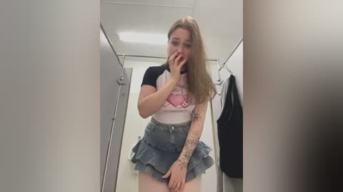 Video of a young woman with long brown hair, wearing a white T-shirt with a pink graphic and a short denim skirt, standing in a public bathroom stall.