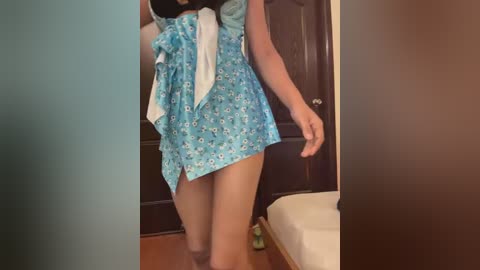 Media: Video of a woman with light skin wearing a blue floral dress with white bows, standing in a room with dark wooden doors and a white cushioned bench.