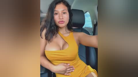 Video of a Latina woman with medium skin tone and long black hair, wearing a yellow one-shoulder dress, sitting in a car, hands on her stomach, with blurred background.