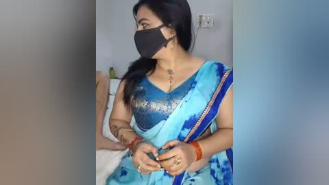 Video of a South Asian woman in a blue and white saree, wearing a black face mask, with a tattoo on her neck, sitting on a bed.