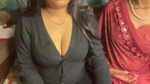 Video of a woman with medium skin tone and long black hair, wearing a deep V-neck ribbed sweater, a pearl necklace, and a red sari with intricate embroidery, seated on a green and red plaid cushion.