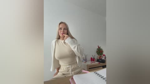 A video of a blonde Caucasian woman in a cream-colored outfit, seated in a minimalist room with a wooden table, notebook, and decorative items.