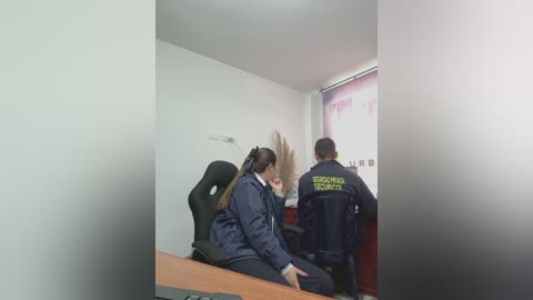 Video of a female officer in navy uniform with a badge, questioning a seated woman in a navy jacket and a ponytail, in a dimly lit, white-walled room with a blurred window and red wall in the background.