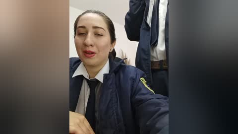 Video of a middle-aged Caucasian woman in a navy blue police uniform with a white shirt and black tie, sitting in a chair, eyes closed, mouth open, with a blurred background.