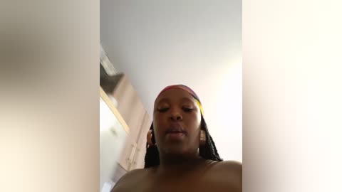 A low-angle, out-of-focus video of a topless black woman with long, braided hair and a red and yellow headscarf, standing in a room with white walls and a ceiling light.