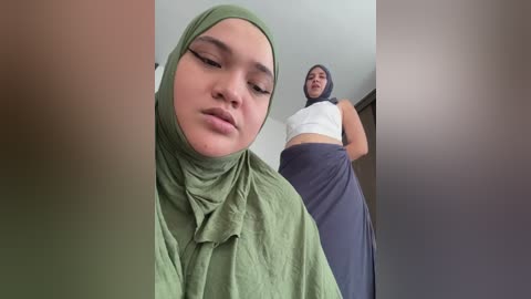 Video of a young woman in a green hijab and a pregnant woman in a white crop top and gray skirt standing behind her.
