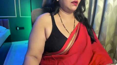 Video of a South Asian woman with long black hair, wearing a red sari with a black blouse, sitting on a chair in a modern, dimly lit room with green and beige curtains.