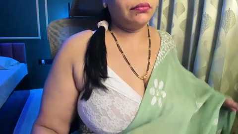 Media: Video of a plus-sized woman with light brown skin, wearing a white lace bra, green floral saree, and gold necklace, seated on a black chair in a modern, dimly lit room with blue curtains.
